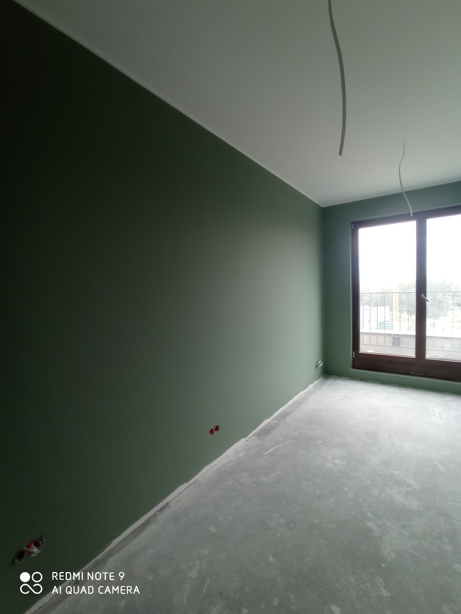 Pomalowane na oliwkowo ściany w pustym pokoju, surowa podłoga, wystające kable z sufitu, okno z brązową ramą.