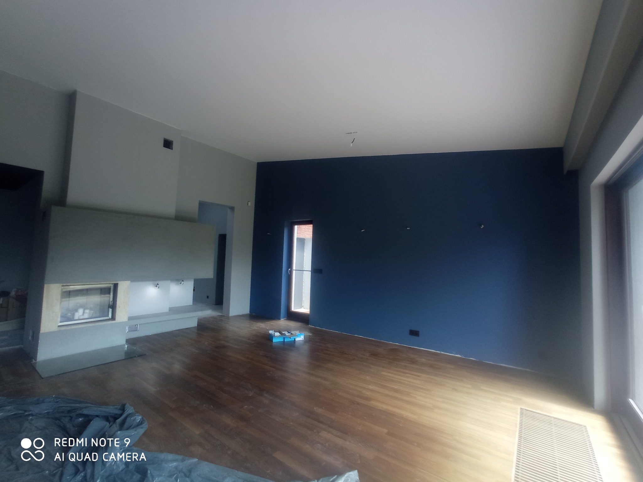 Wnętrze pokoju po remoncie z pomalowaną na granatowo ścianą, drewnianą podłogą, nowoczesnym kominkiem i oknem balkonowym. Na podłodze widoczne narzędzia malarskie.