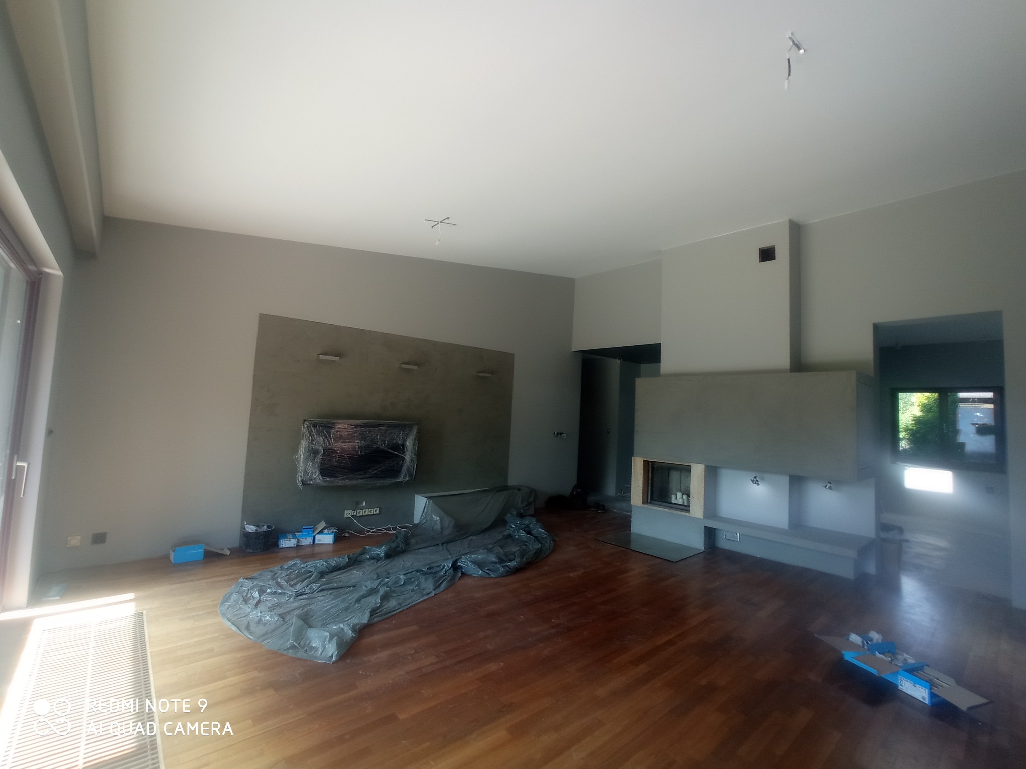 Pokój w trakcie remontu z drewnianą podłogą, szarymi ścianami, osłoniętym folią telewizorem i zabudowanym kominkiem, widoczne materiały budowlane.