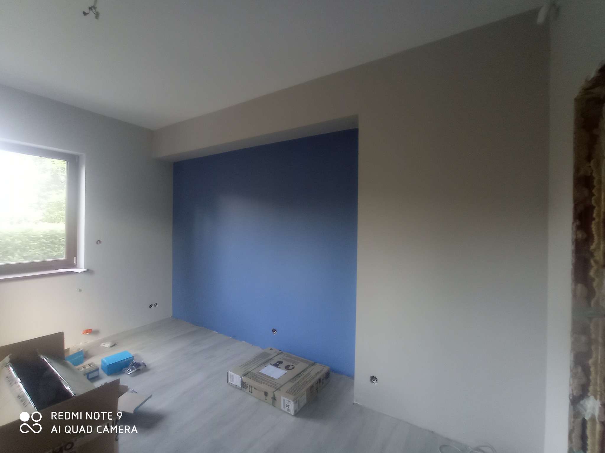 Wnętrze pokoju w trakcie prac wykończeniowych: pomalowana na niebiesko wnęka, jasne ściany, częściowo położona podłoga, widoczne puszki elektryczne i narzędzia w kartonie.