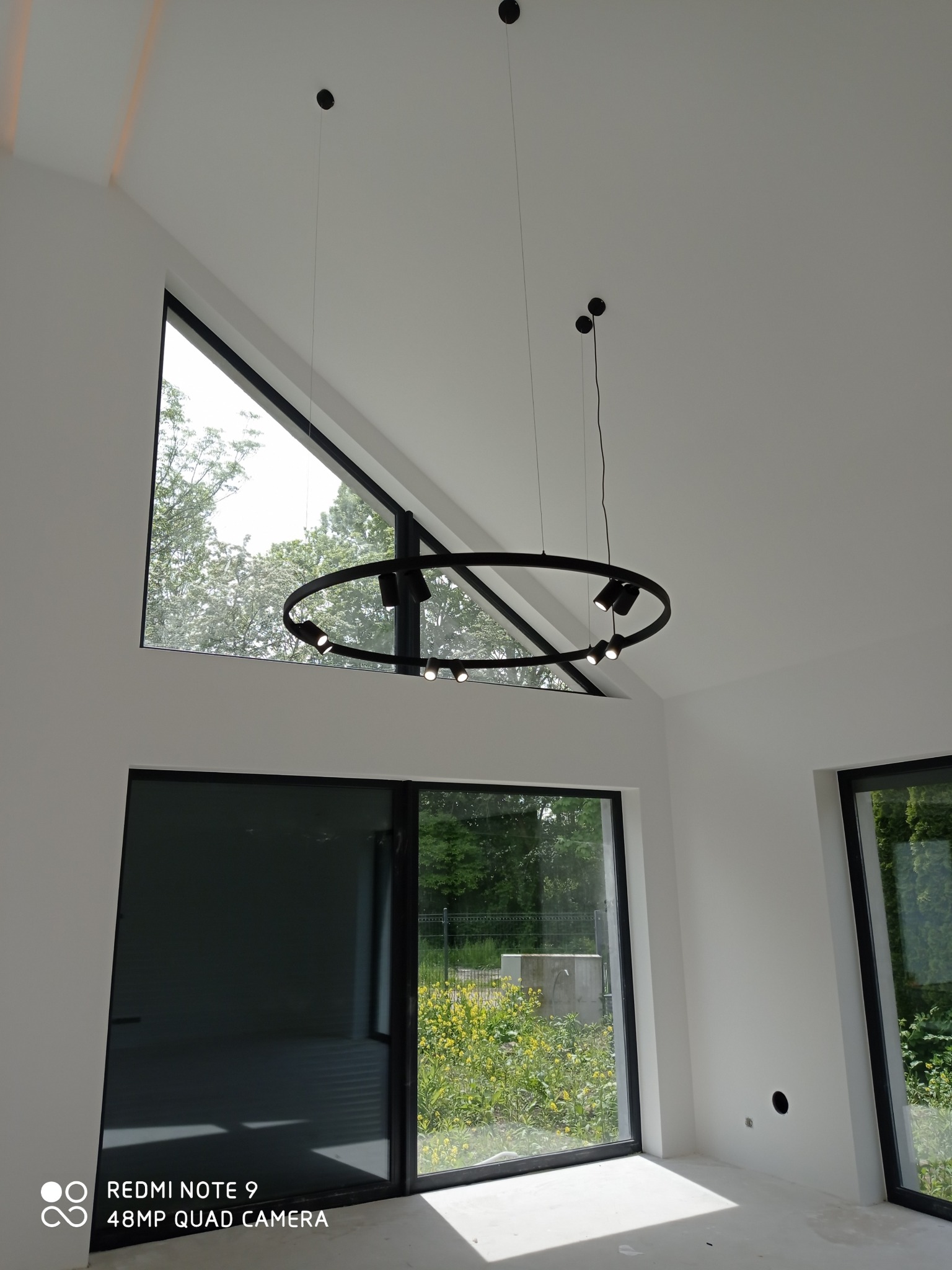 Wnętrze z wysokim stropem i trójkątnym oknem, nowoczesna okrągła lampa wisząca z reflektorami, duże przesuwne drzwi balkonowe z widokiem na zieleń, surowe wykończenie podłogi.