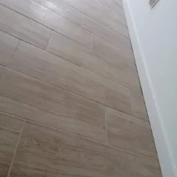 Ułożone pod kątem płytki podłogowe imitujące drewno, przylegające do białej ściany z gniazdkiem elektrycznym.