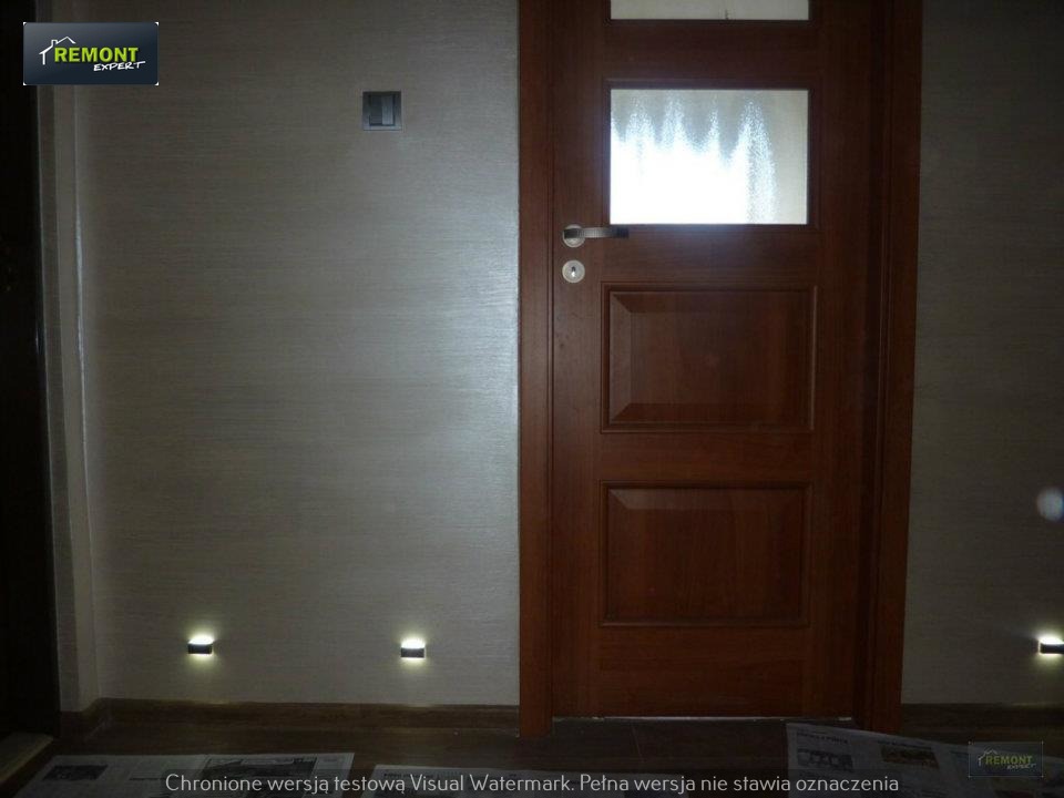 Wykończone wnętrze korytarza z drewnianymi drzwiami z szybą, gładką ścianą w odcieniach beżu i małymi punktowymi lampkami LED przy podłodze. Widoczny fragment gazety na podłodze.