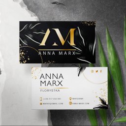Oliwia Koligot - Dwie wizytówki z motywem florystycznym, jedna czarna z złotym logo AM i liśćmi, druga biała z danymi kontaktowymi florystki Anny Marx, ułożone na tle z szarego kamienia i zielonych liści palmowych.