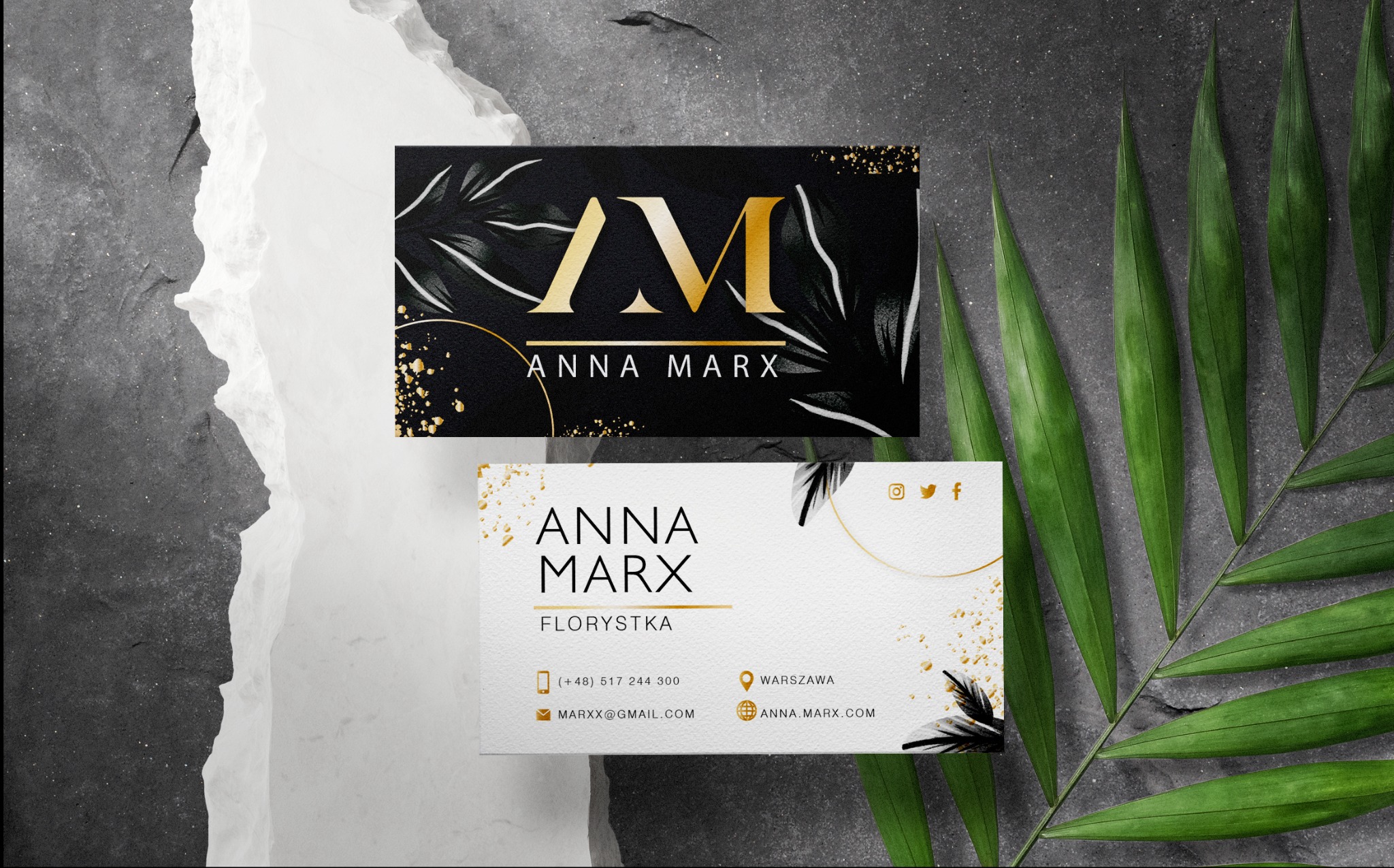 Dwie wizytówki z motywem florystycznym, jedna czarna z złotym logo AM i liśćmi, druga biała z danymi kontaktowymi florystki Anny Marx, ułożone na tle z szarego kamienia i zielonych liści palmowych.