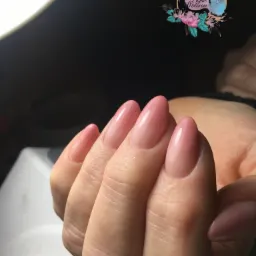 Dłoń z pomalowanymi paznokciami w odcieniach różu, z widocznym logo salonu w górnym rogu i lampą UV w tle.