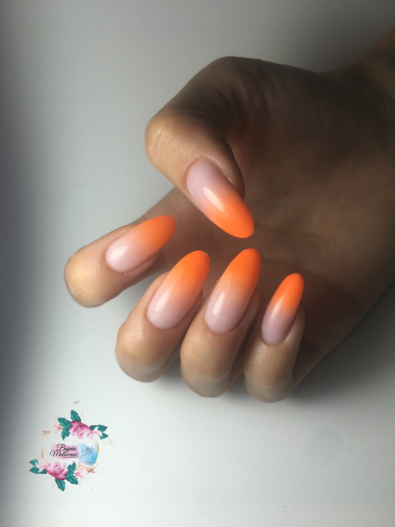 Dłoń z długimi, owalnymi paznokciami z efektem ombre: od jasnoróżowego u nasady do intensywnego pomarańczowego na końcach. Logo salonu w rogu.