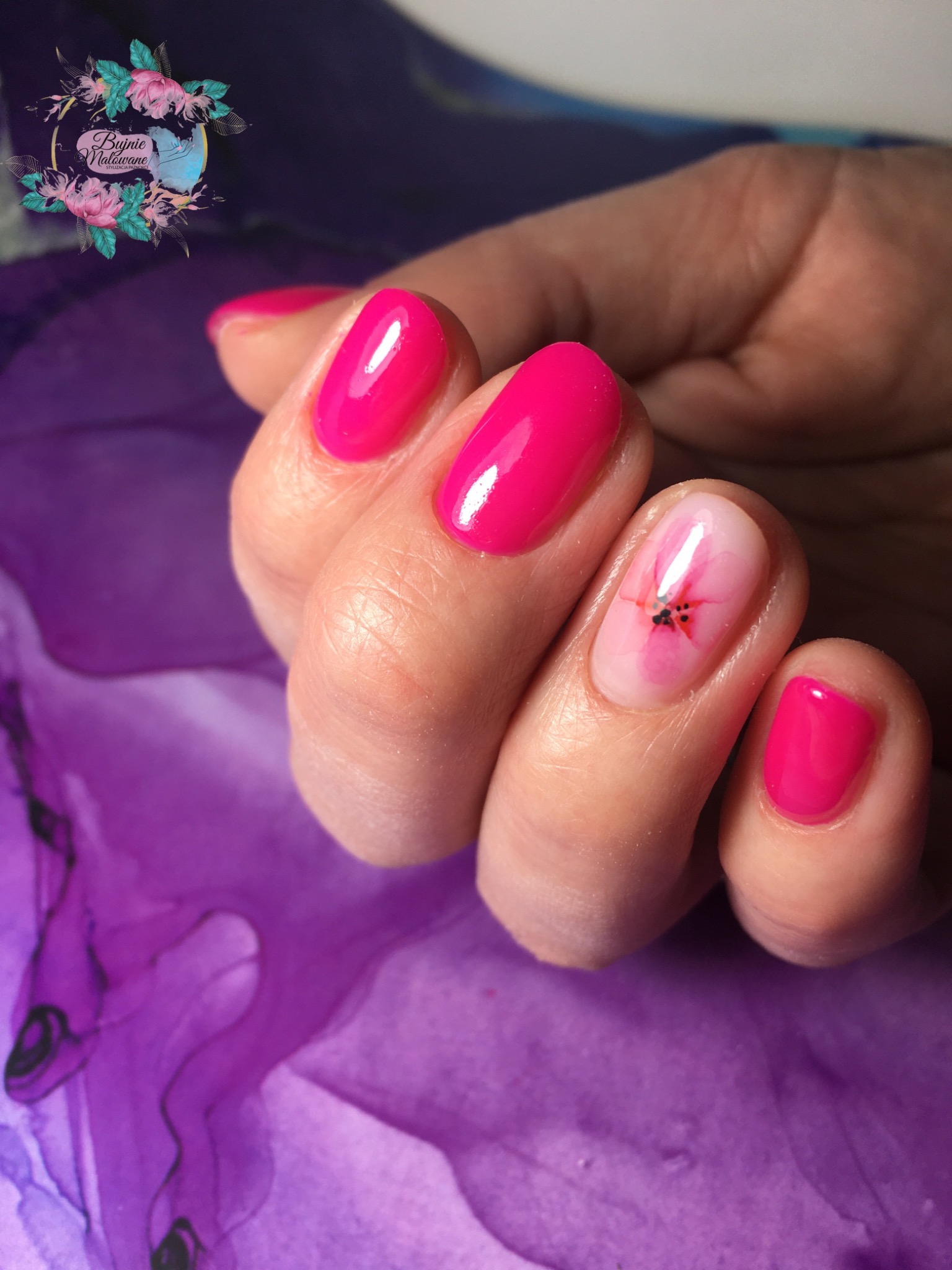Dłoń z pomalowanymi paznokciami w kolorze fuksji, jeden paznokieć z delikatnym kwiatowym wzorem, na rozmytym fioletowym tle.
