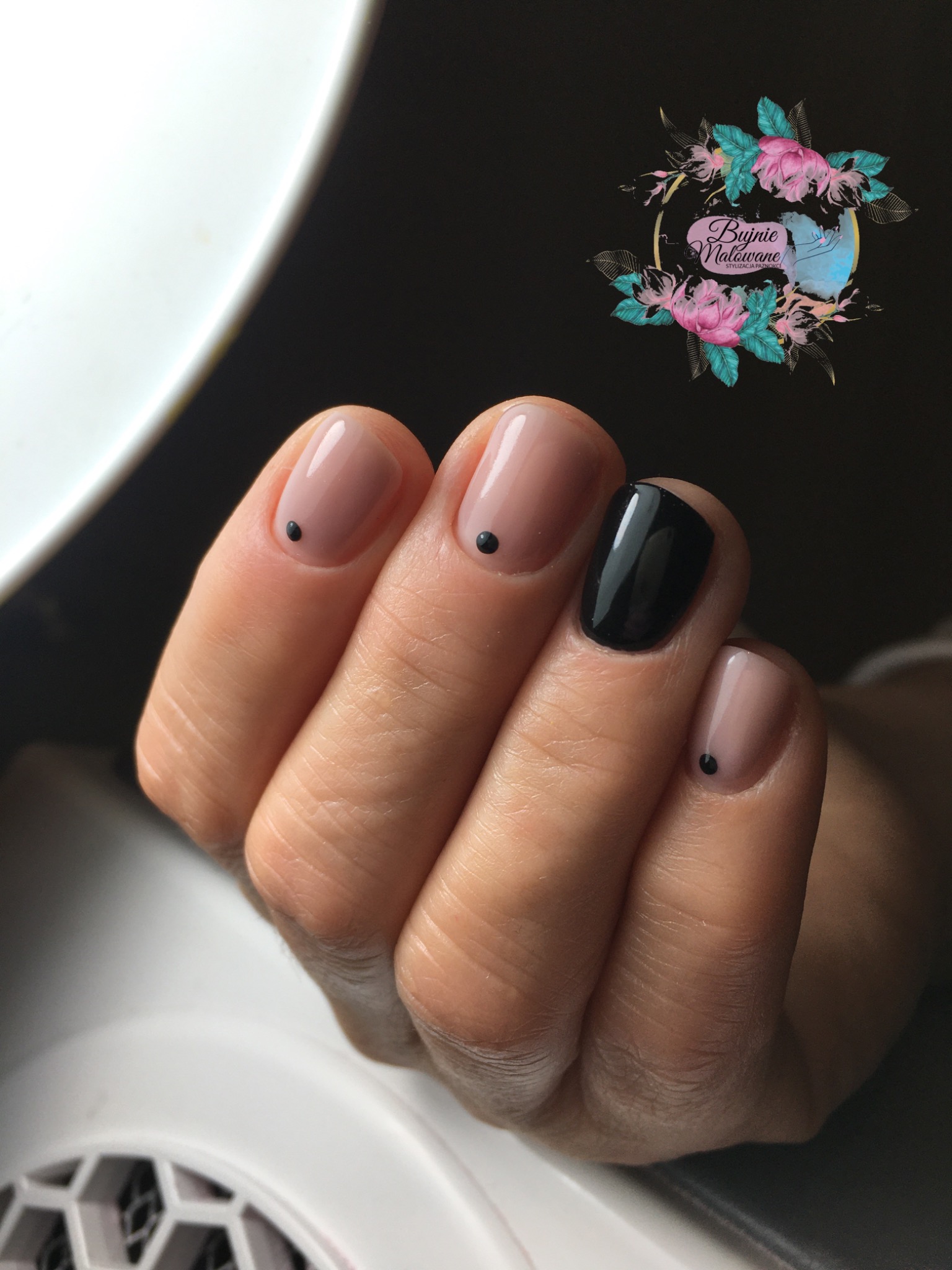 Dłoń z minimalistycznym zdobieniem paznokci: trzy paznokcie w odcieniu nude z pojedynczą czarną kropką u nasady, jeden paznokieć pomalowany na czarno na wysoki połysk. W tle widoczne logo salonu...