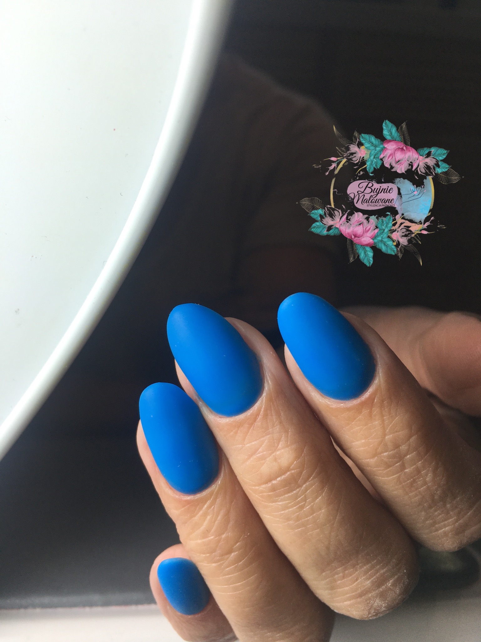 Matowe, niebieskie paznokcie w kształcie migdała, na tle lampy do utwardzania i logo salonu stylizacji paznokci z motywem kwiatowym.