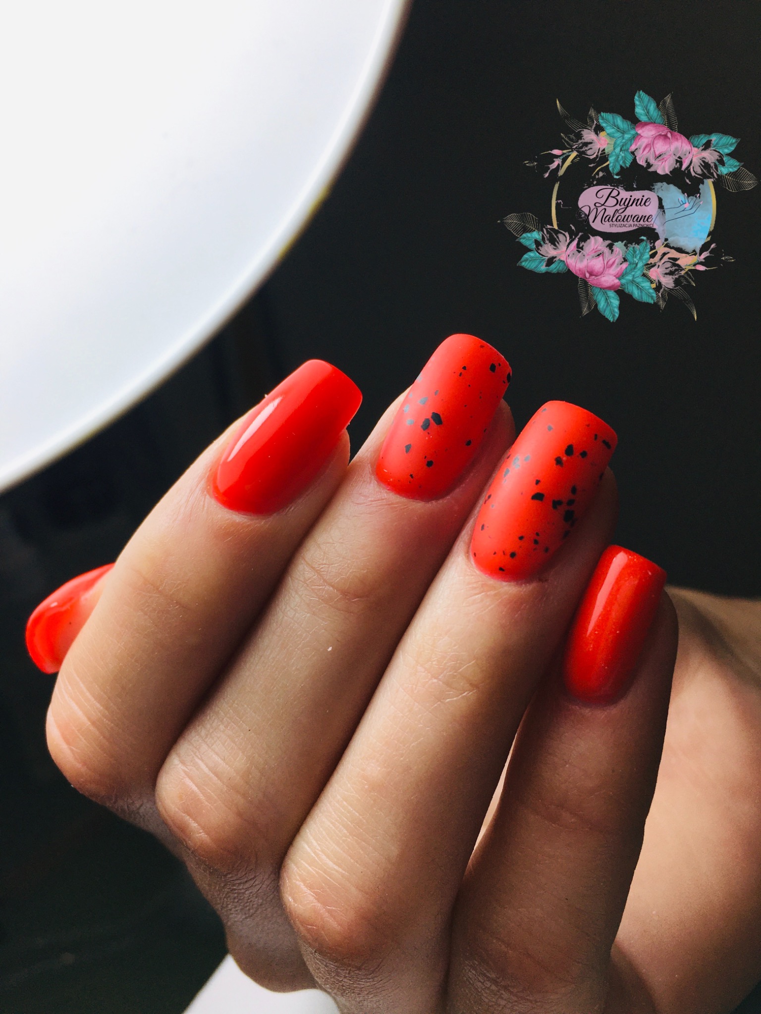 Pomarańczowe paznokcie w geometryczne czarne kropki, dwa z nich z błyszczącym wykończeniem, oświetlone lampą pierścieniową, logo salonu w tle.