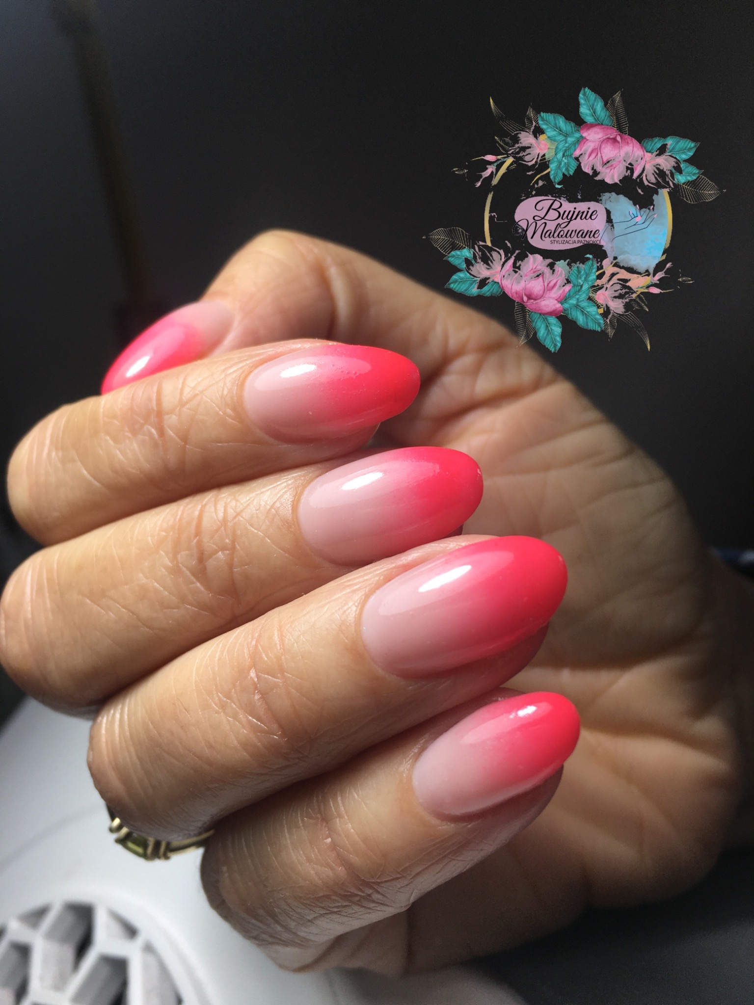 Dłoń z owalnymi paznokciami pomalowanymi lakierem w odcieniach różu, tworzącym efekt ombre, z pierścionkiem na palcu, na rozmytym tle z logo salonu.