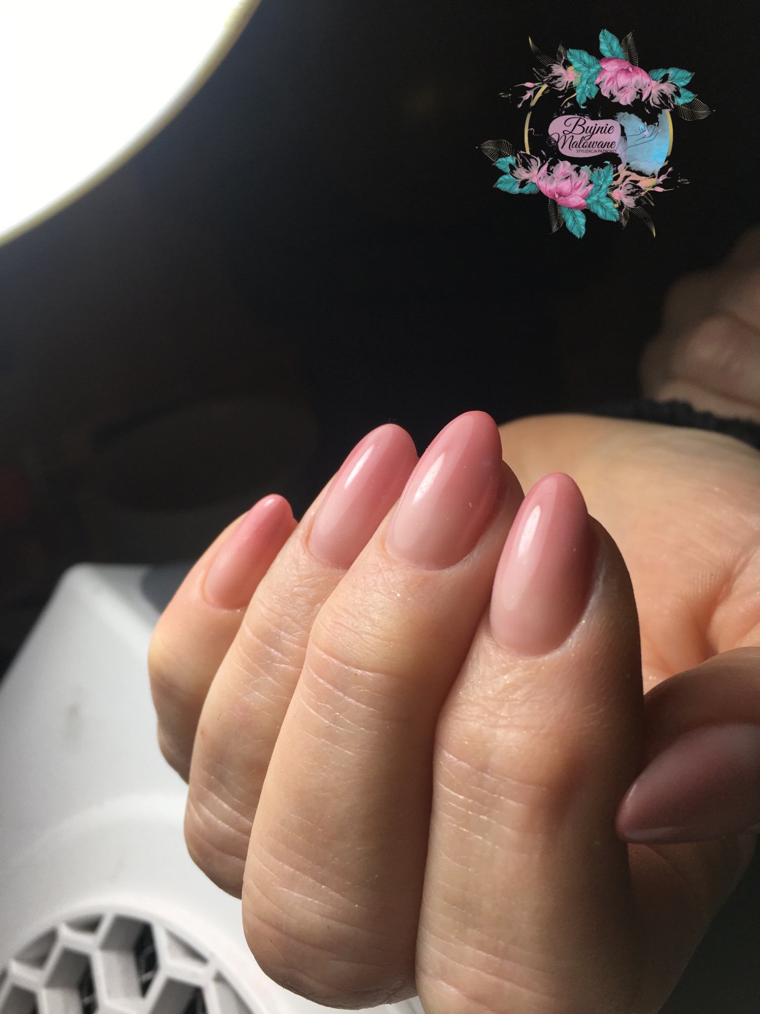 Dłoń z pomalowanymi paznokciami w odcieniach różu, z widocznym logo salonu w górnym rogu i lampą UV w tle.