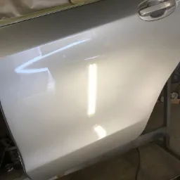 Świeżo polakierowane, srebrne drzwi samochodu z widoczną klamką, zabezpieczone taśmą i papierem przed dalszymi pracami blacharsko-lakierniczymi, ustawione na metalowym stelażu.