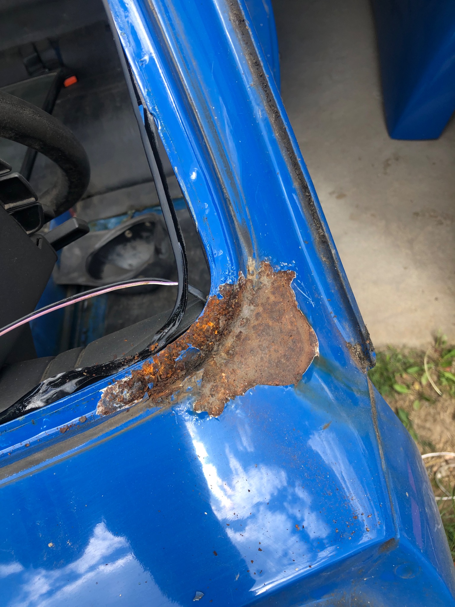 Fragment niebieskiego nadwozia pojazdu z widoczną, rozległą korozją w okolicy ramy okna, z widocznymi fragmentami rdzy i łuszczącej się farby.