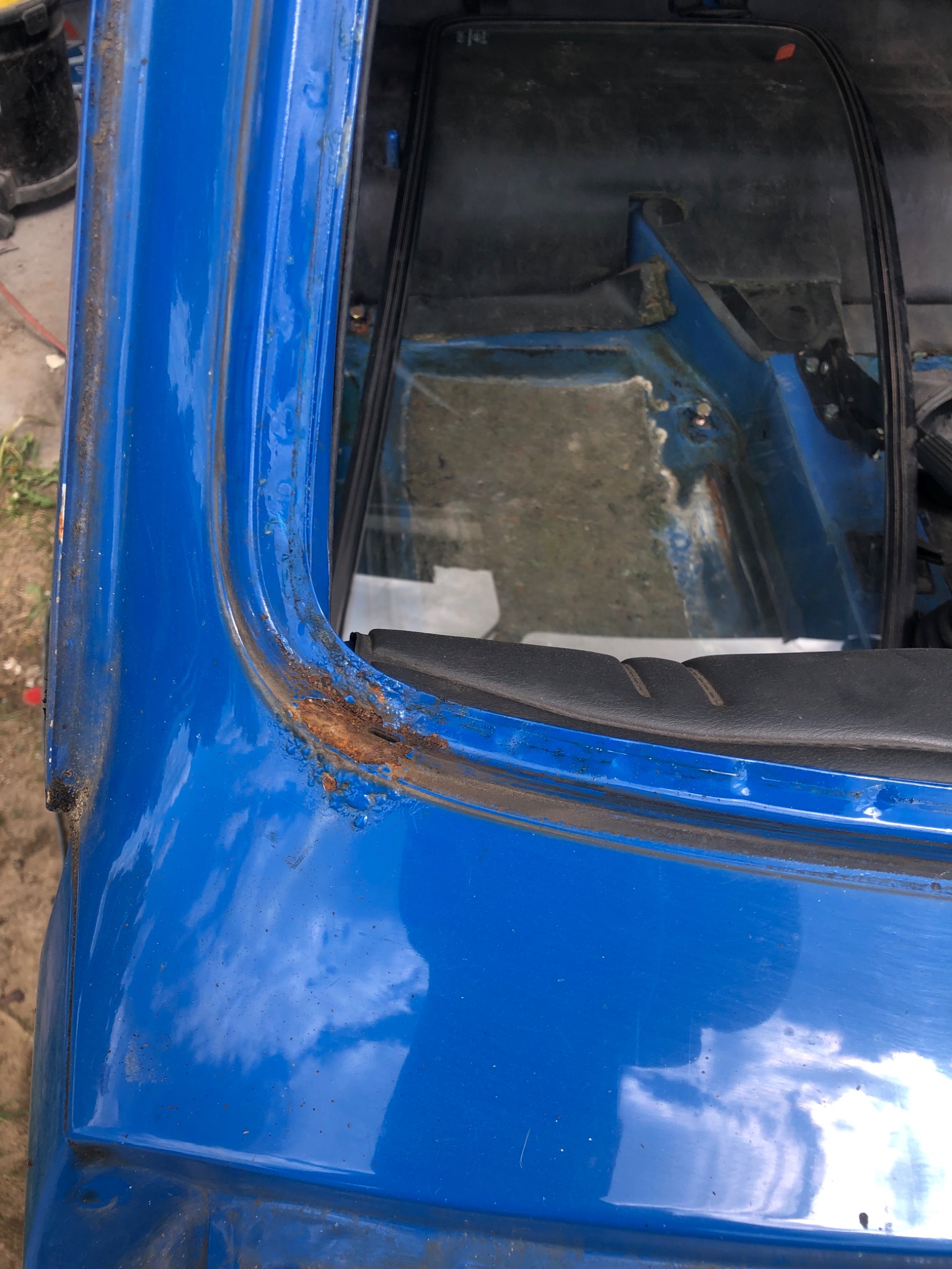 Fragment niebieskiego nadwozia samochodu z widoczną korozją wokół ramy okna, widoczne ślady rdzy i odpryski lakieru.