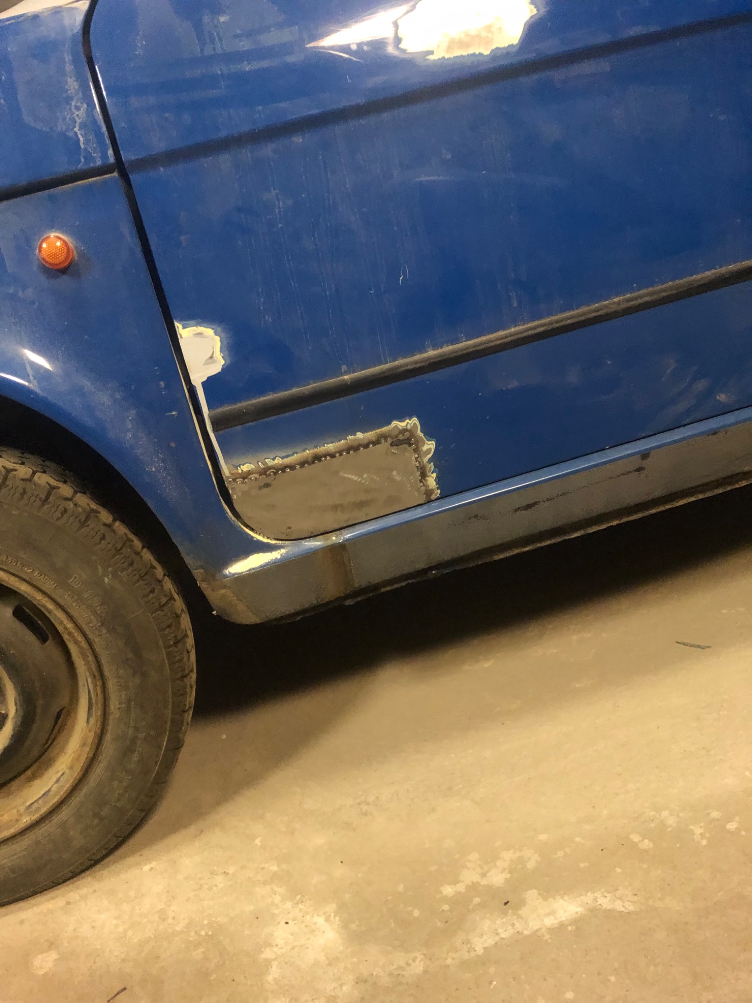 Fragment niebieskiego samochodu w trakcie naprawy blacharskiej, widoczne świeże spawy i obszary przygotowane do lakierowania, zarys opony w lewym dolnym rogu.