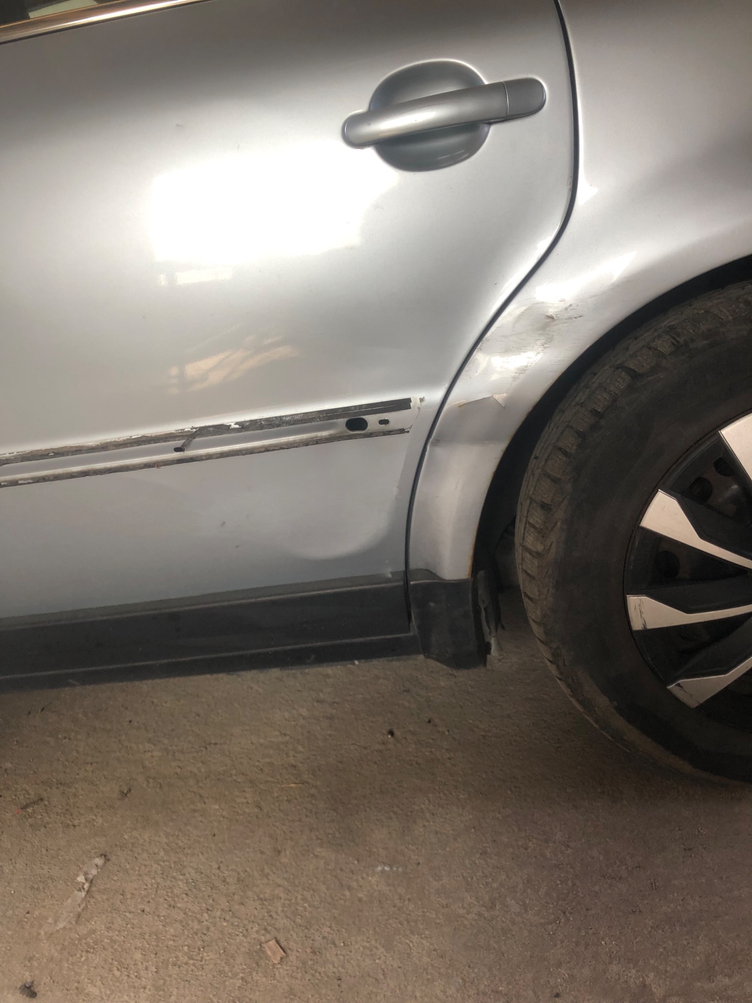 Uszkodzony srebrny samochód: wgniecenie na drzwiach, brak listwy ochronnej, widoczna rdza przy nadkolu, opona z czarną felgą z białymi elementami, betonowa podłoga.