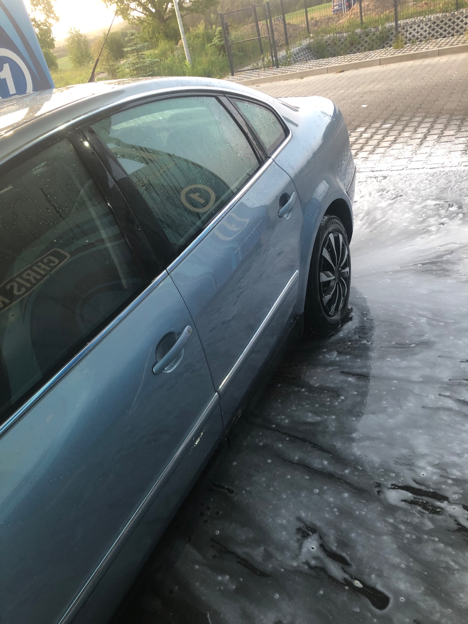 Jasnoniebieski samochód osobowy z czarnymi felgami, myty pianą aktywną w myjni bezdotykowej, widok z boku na przednie drzwi i koło, w tle zieleń drzew.