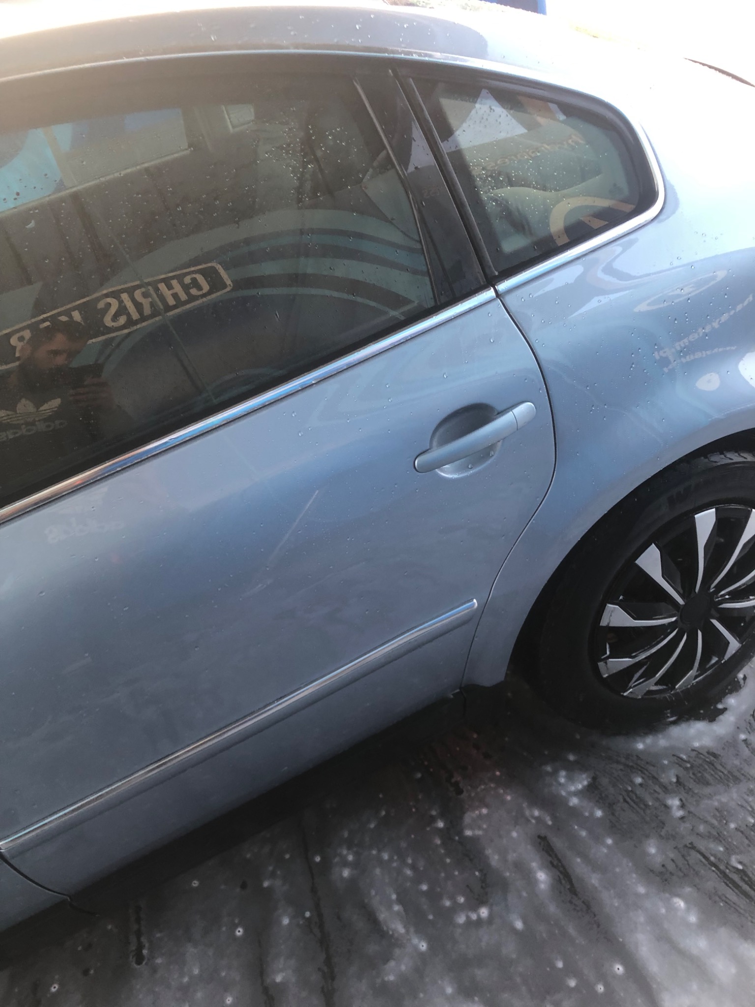 Błękitny samochód osobowy, po umyciu, z widocznymi kroplami wody na karoserii i pianą na ziemi, felgi aluminiowe z czarno-srebrnym wzorem, w odbiciu w szybie widoczna osoba w bluzie marki Adidas.