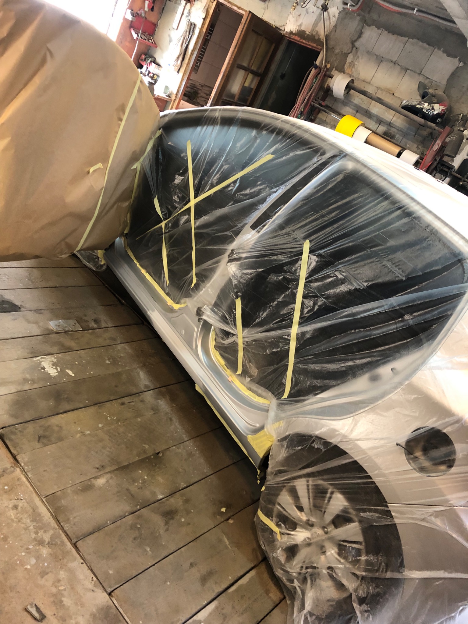 Samochód osobowy przygotowany do lakierowania, osłonięty folią i papierem maskującym, z widocznymi taśmami zabezpieczającymi, w warsztacie z drewnianą podłogą.