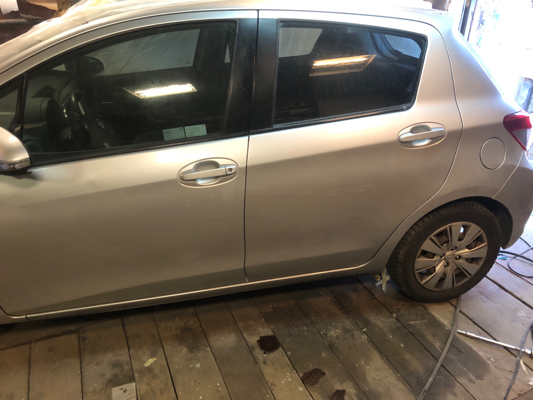 Srebrny samochód Toyota Yaris, widok z boku, zaparkowany w warsztacie na drewnianej podłodze. Widoczne zaklejone taśmą fragmenty podwozia.