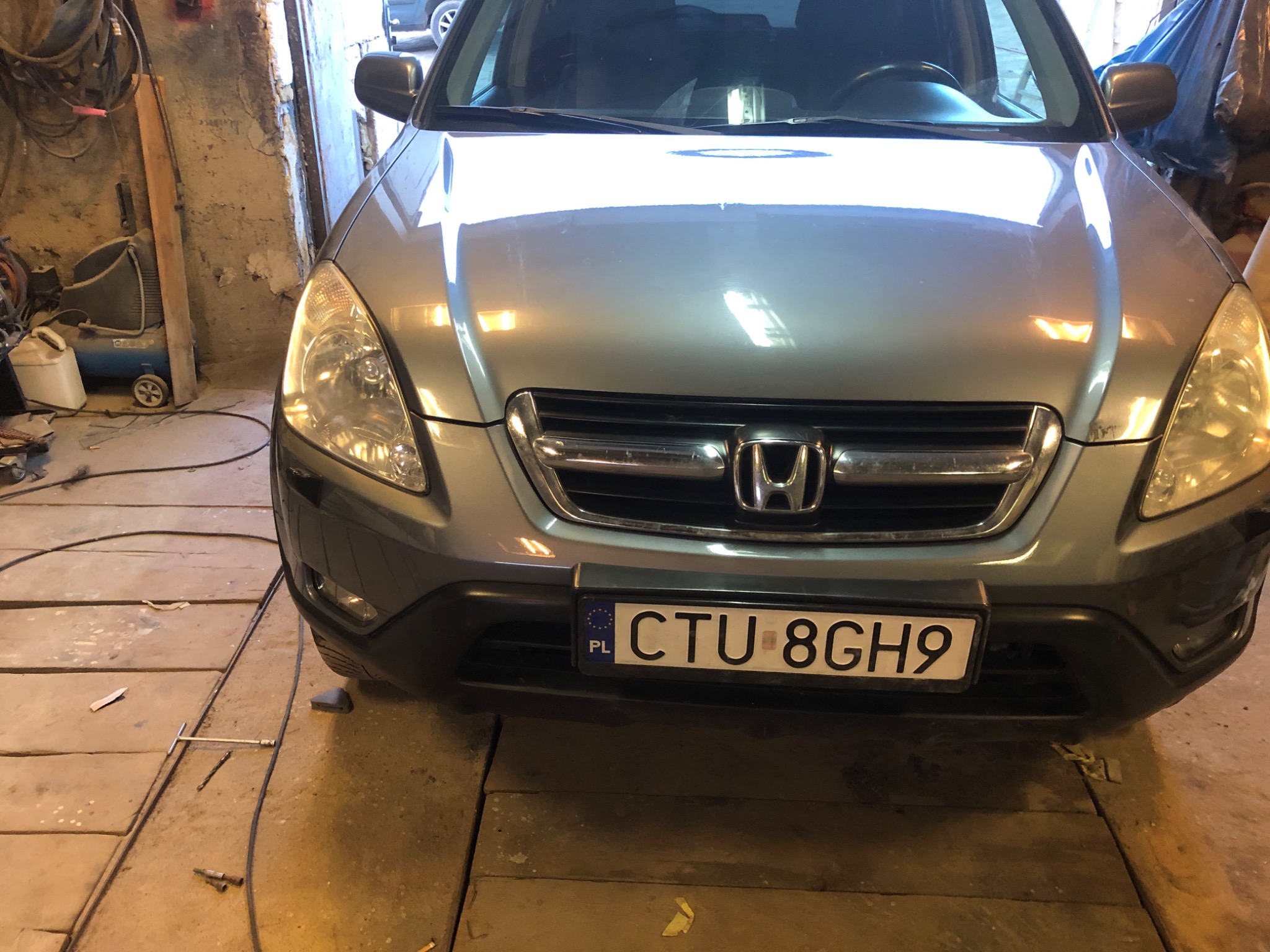 Przód srebrnego samochodu Honda CR-V z polską tablicą rejestracyjną, widoczny w warsztacie z betonową ścianą i narzędziami na podłodze.