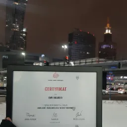 Oprawiony certyfikat ukończenia szkolenia z zakresu mobilności, stabilności i wzorców ruchowych dla Ewy Kalickiej, na tle nocnego krajobrazu Warszawy z Pałacem Kultury i Nauki, trzymany w ręce...