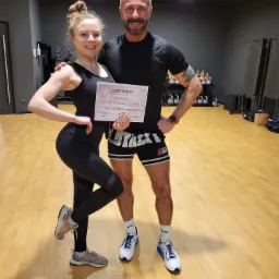 Kobieta w sportowym stroju pozuje z certyfikatem PFS Full Body Workout obok mężczyzny w sali treningowej z ciężarkami w tle.