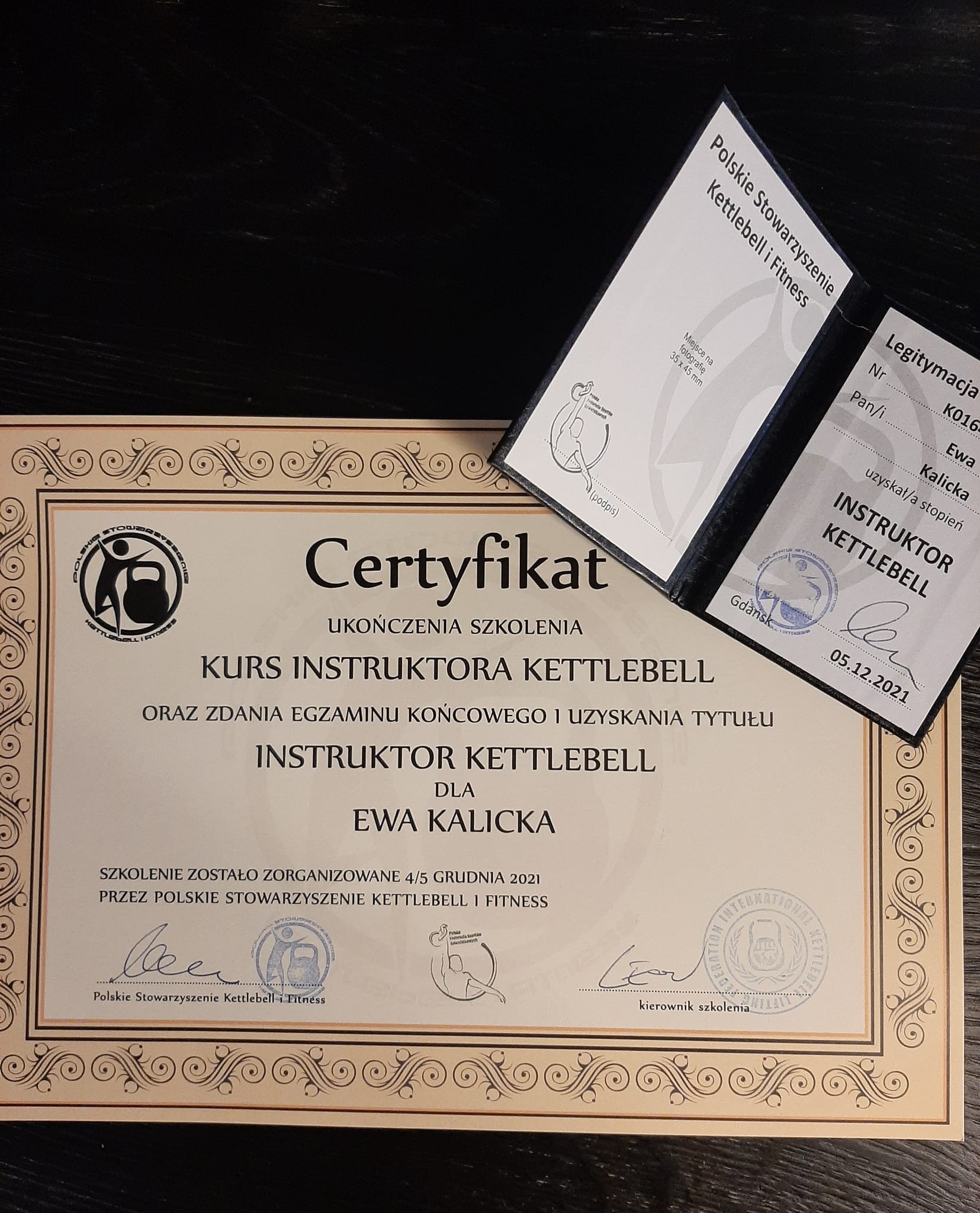 Certyfikat ukończenia kursu instruktora kettlebell oraz legitymacja Polskiego Stowarzyszenia Kettlebell i Fitness, leżące na ciemnym, drewnianym podłożu.