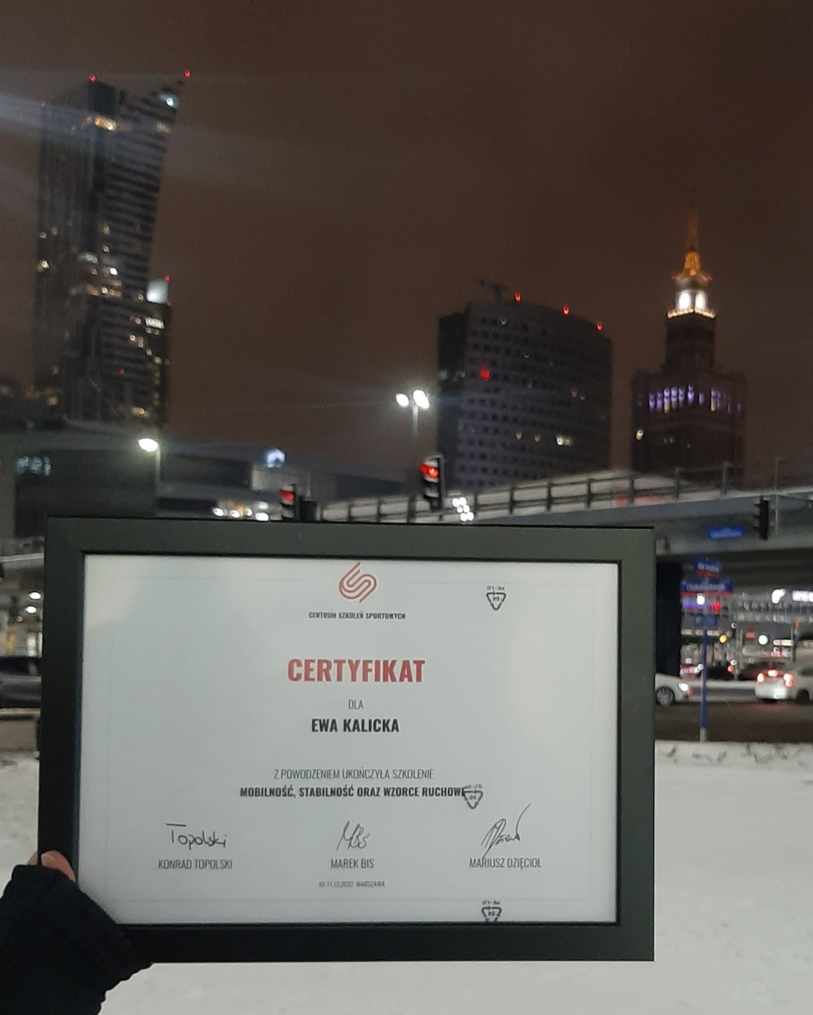 Oprawiony certyfikat ukończenia szkolenia z zakresu mobilności, stabilności i wzorców ruchowych dla Ewy Kalickiej, na tle nocnego krajobrazu Warszawy z Pałacem Kultury i Nauki, trzymany w ręce...