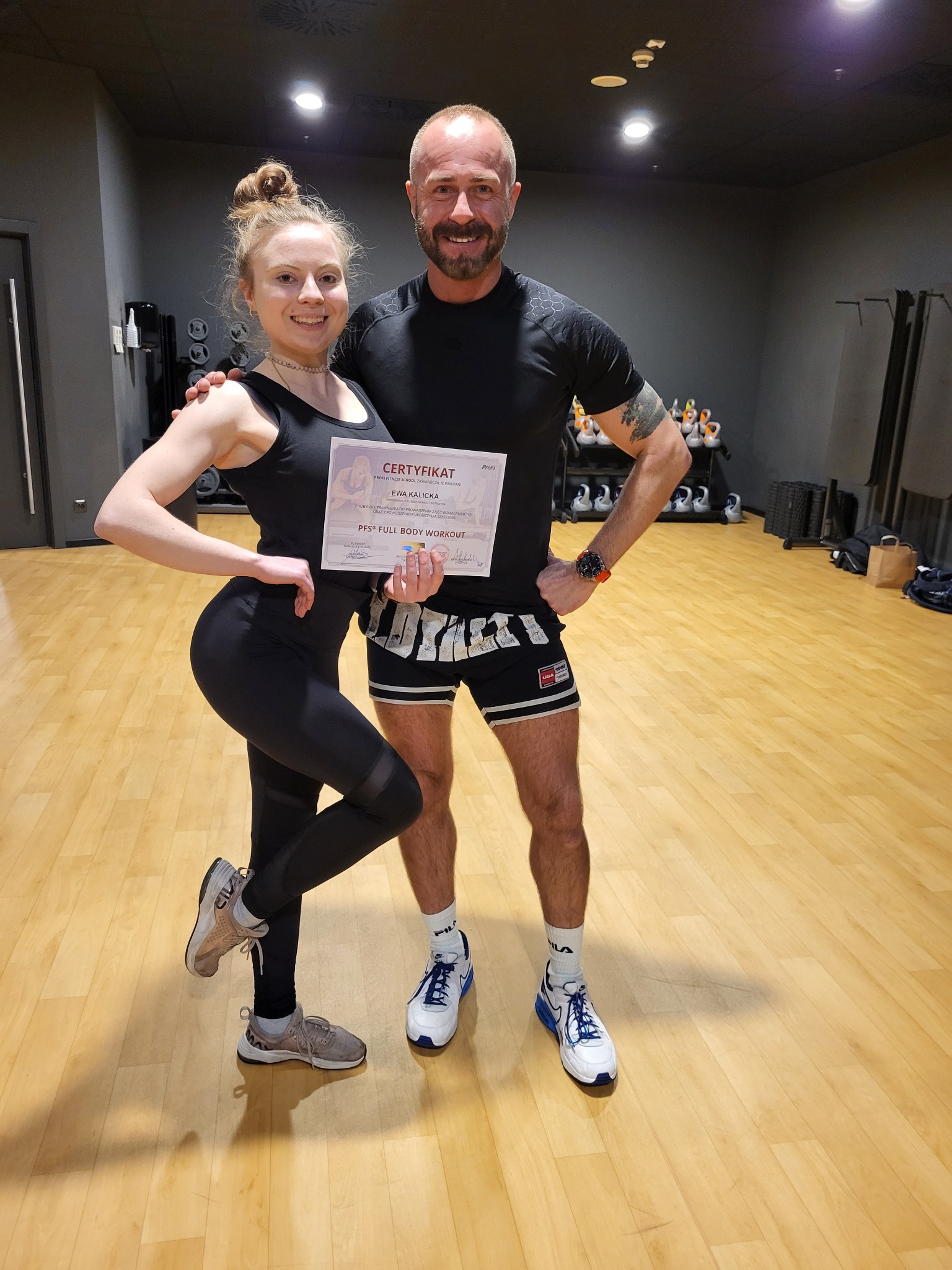 Kobieta w sportowym stroju pozuje z certyfikatem PFS Full Body Workout obok mężczyzny w sali treningowej z ciężarkami w tle.