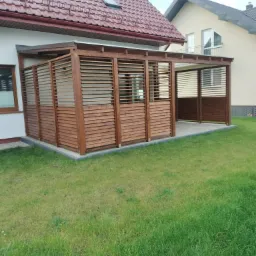 Drewniana pergola z poziomymi listwami, przylegająca do domu z czerwonym dachem, otoczona trawnikiem.