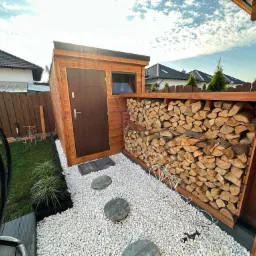 Drewniana drewutnia z zadaszeniem i drzwiami, wypełniona pociętym drewnem opałowym, otoczona białym żwirem i kamiennymi płytami, widok z niskiej perspektywy w słoneczny dzień.