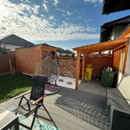 Nowoczesny taras z desek kompozytowych w odcieniach szarości, z drewnianą pergolą, miejscem na składowanie drewna i wiszącym fotelem, uzupełniony elementami małej architektury ogrodowej.