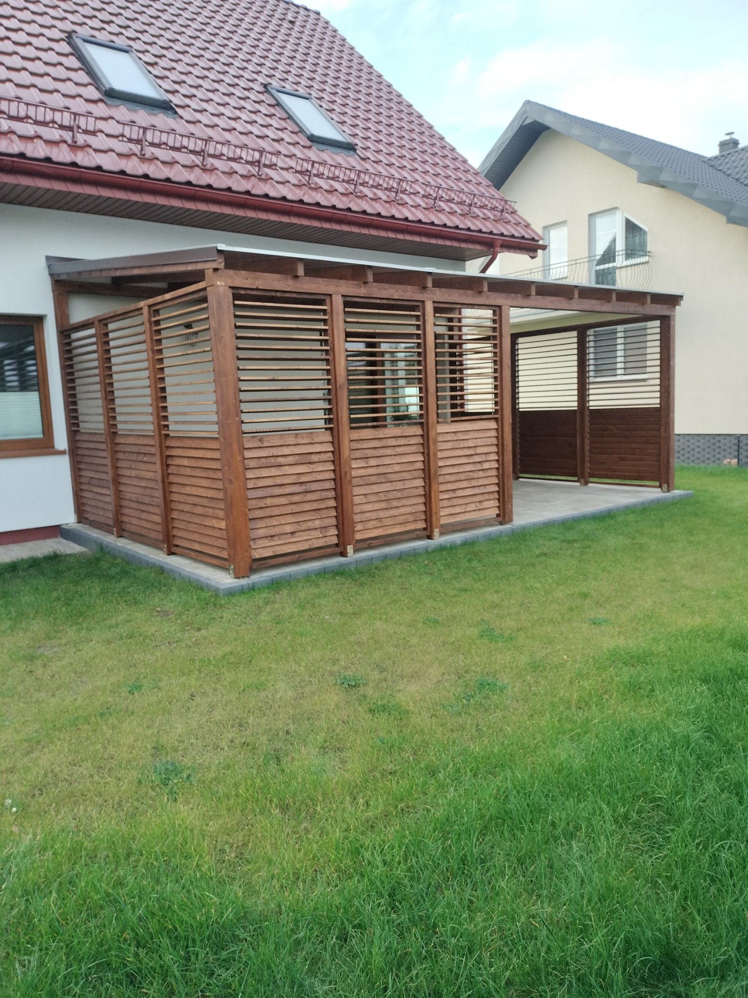 Drewniana pergola z poziomymi listwami, przylegająca do domu z czerwonym dachem, otoczona trawnikiem.
