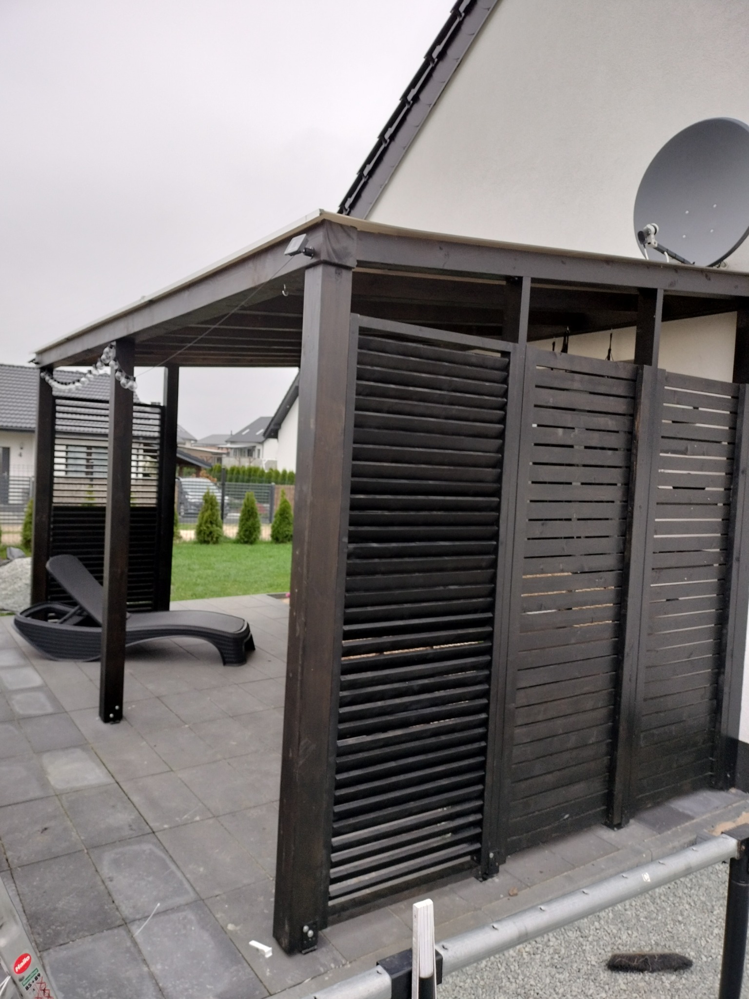 Ciemna, drewniana pergola z żaluzjowymi ścianami, na szarym brukowanym tarasie, z leżakiem w środku, oświetlona girlandą żarówek, z satelitą na dachu domu w tle.