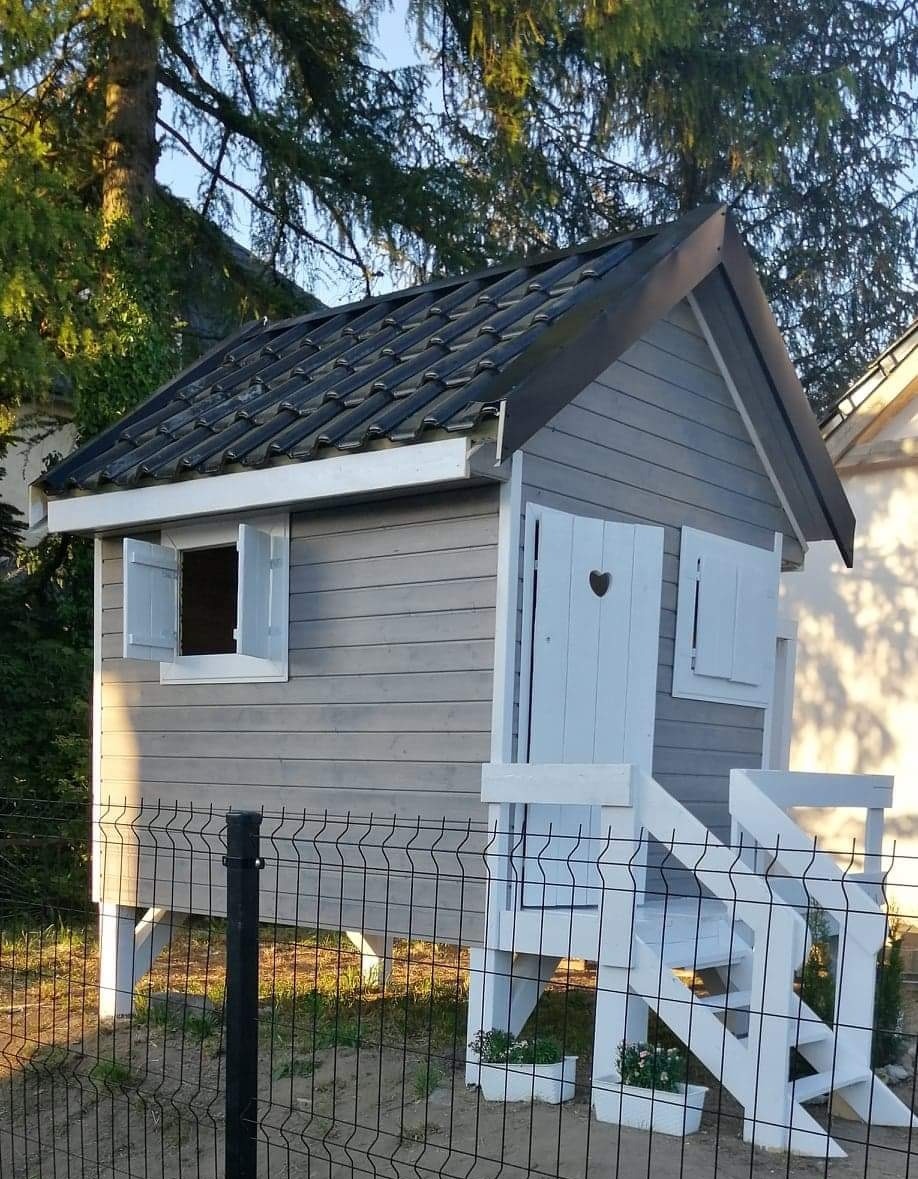 Szary drewniany domek dla dzieci z czarnym dachem z dachówki, białymi okiennicami i schodkami, widoczny za czarnym metalowym ogrodzeniem.