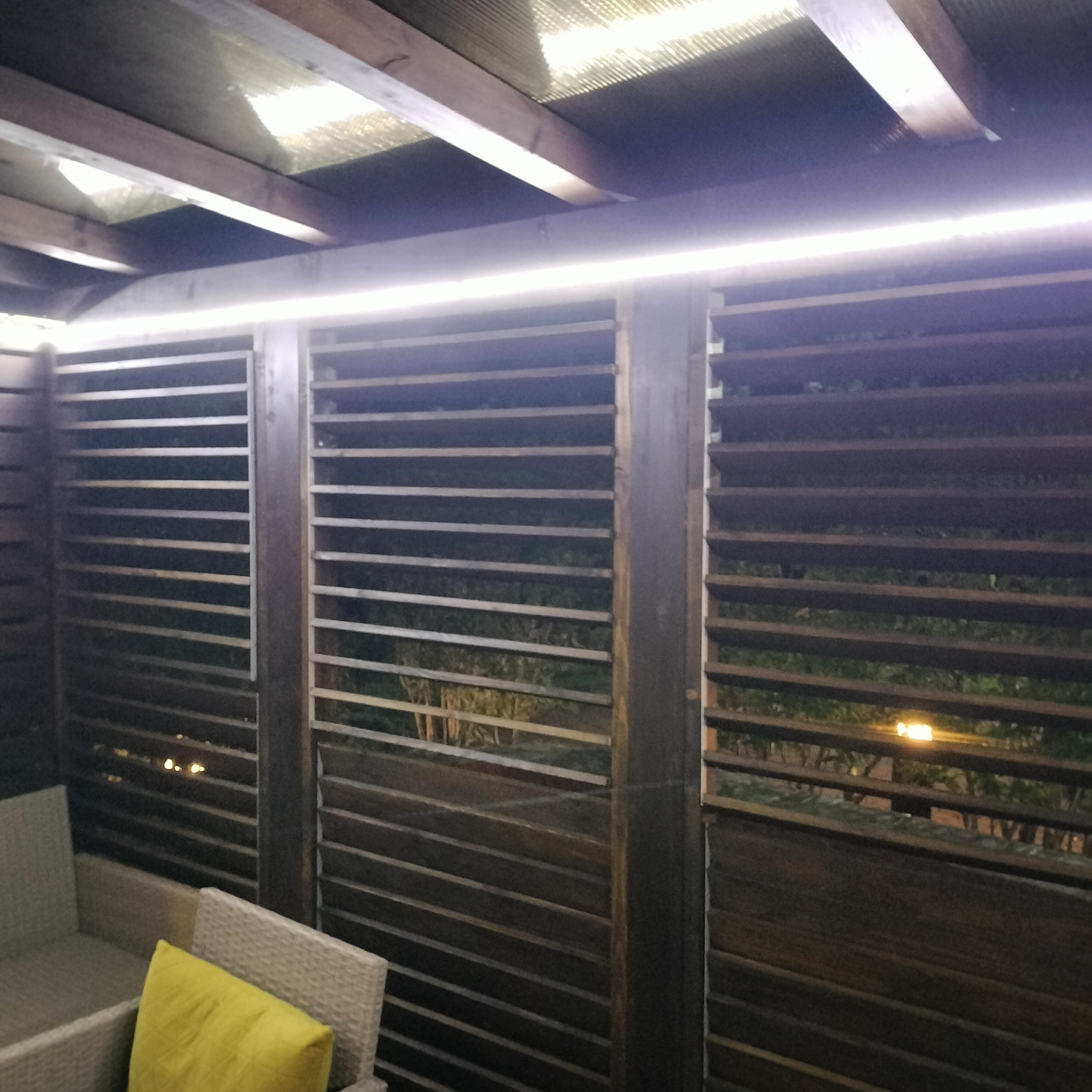 Zadaszona konstrukcja z drewnianymi lamelami, oświetlona listwą LED, z widocznym fragmentem mebli ogrodowych z żółtą poduszką.