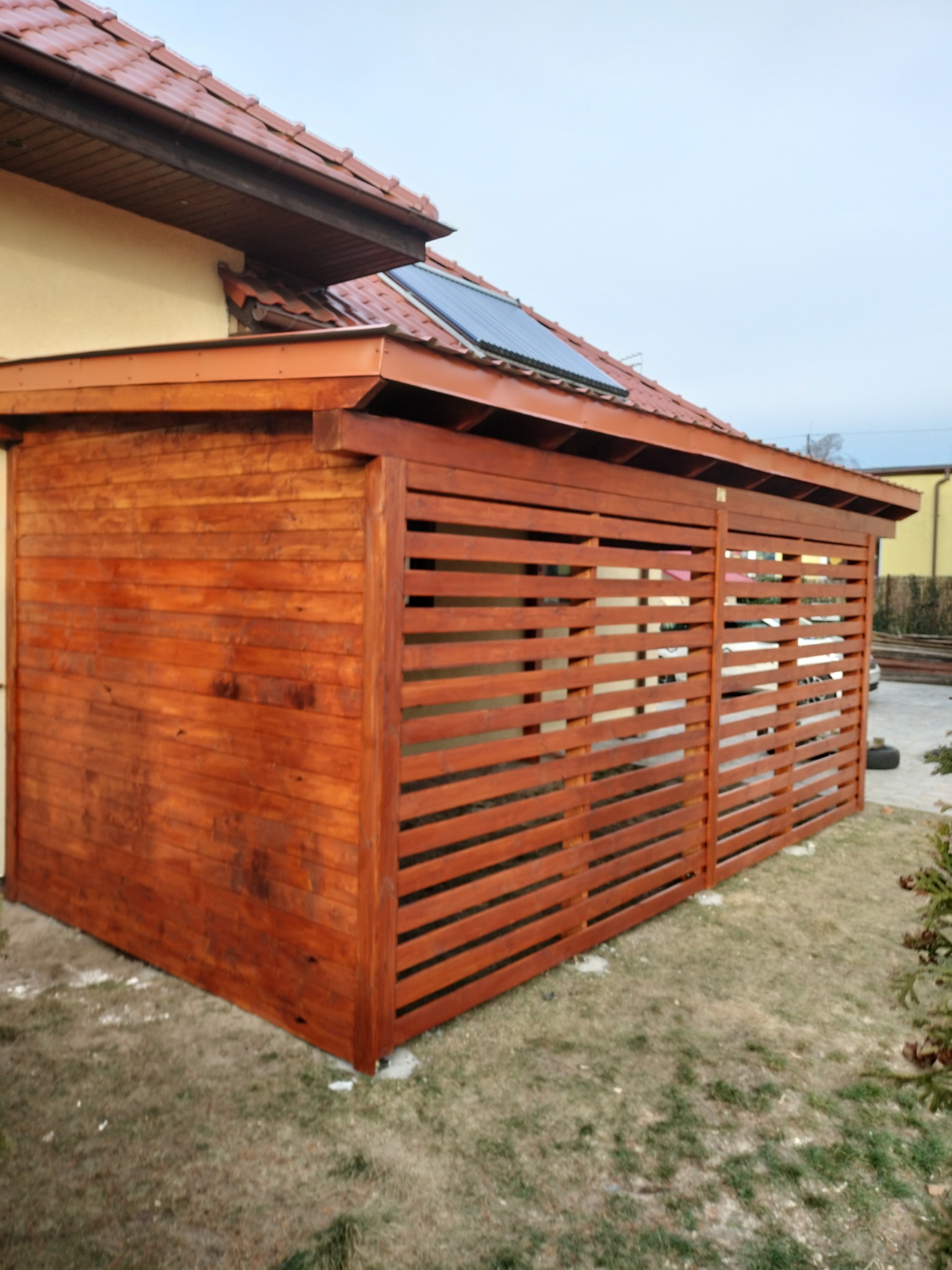 Drewniana konstrukcja z poziomych listew, przylegająca do budynku z brązowym dachem, widoczna z zewnątrz, na trawiastym terenie.