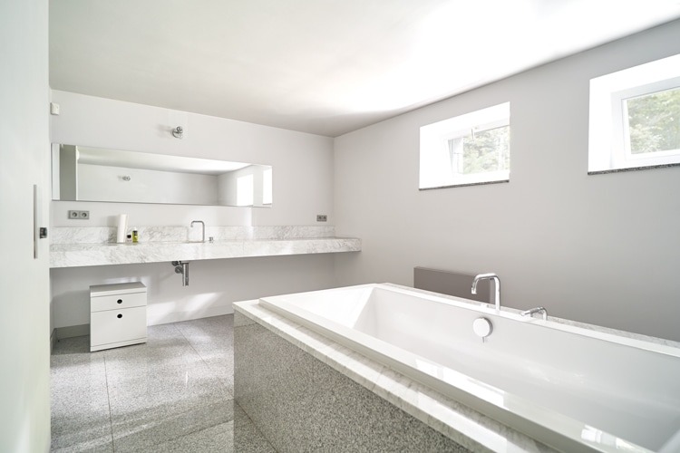 Minimalistyczna łazienka w odcieniach szarości i bieli, z wanną obudowaną szarym kamieniem, długim blatem z umywalkami i dużym lustrem, oraz dwoma oknami wpuszczającymi naturalne światło.