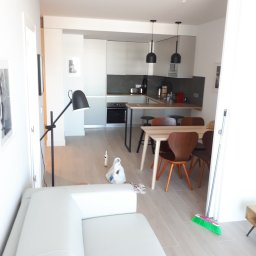 Paweł Plashenko - Jasne, nowoczesne wnętrze mieszkania z otwartą kuchnią i salonem, widoczny biały narożnik, stół z krzesłami, lampa stojąca, oraz akcesoria malarskie i miotła w rogu, co sugeruje prace wykończeniowe.