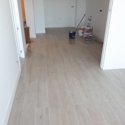 Paweł Plashenko - Ułożone jasne płytki podłogowe imitujące drewno w nowym mieszkaniu, widoczne białe ściany i listwy przypodłogowe, wiadro, mop i odkurzacz przemysłowy w tle.