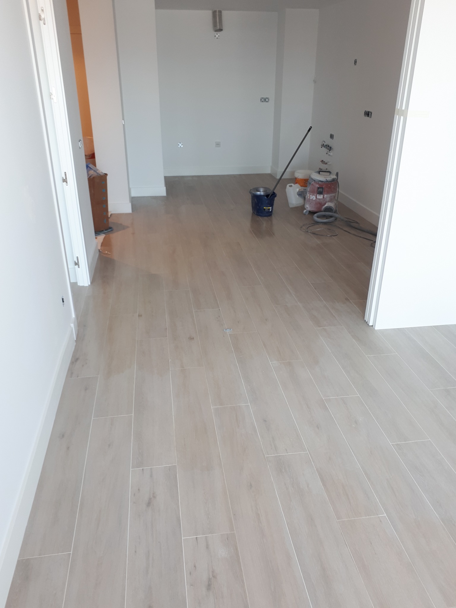 Ułożone jasne płytki podłogowe imitujące drewno w nowym mieszkaniu, widoczne białe ściany i listwy przypodłogowe, wiadro, mop i odkurzacz przemysłowy w tle.