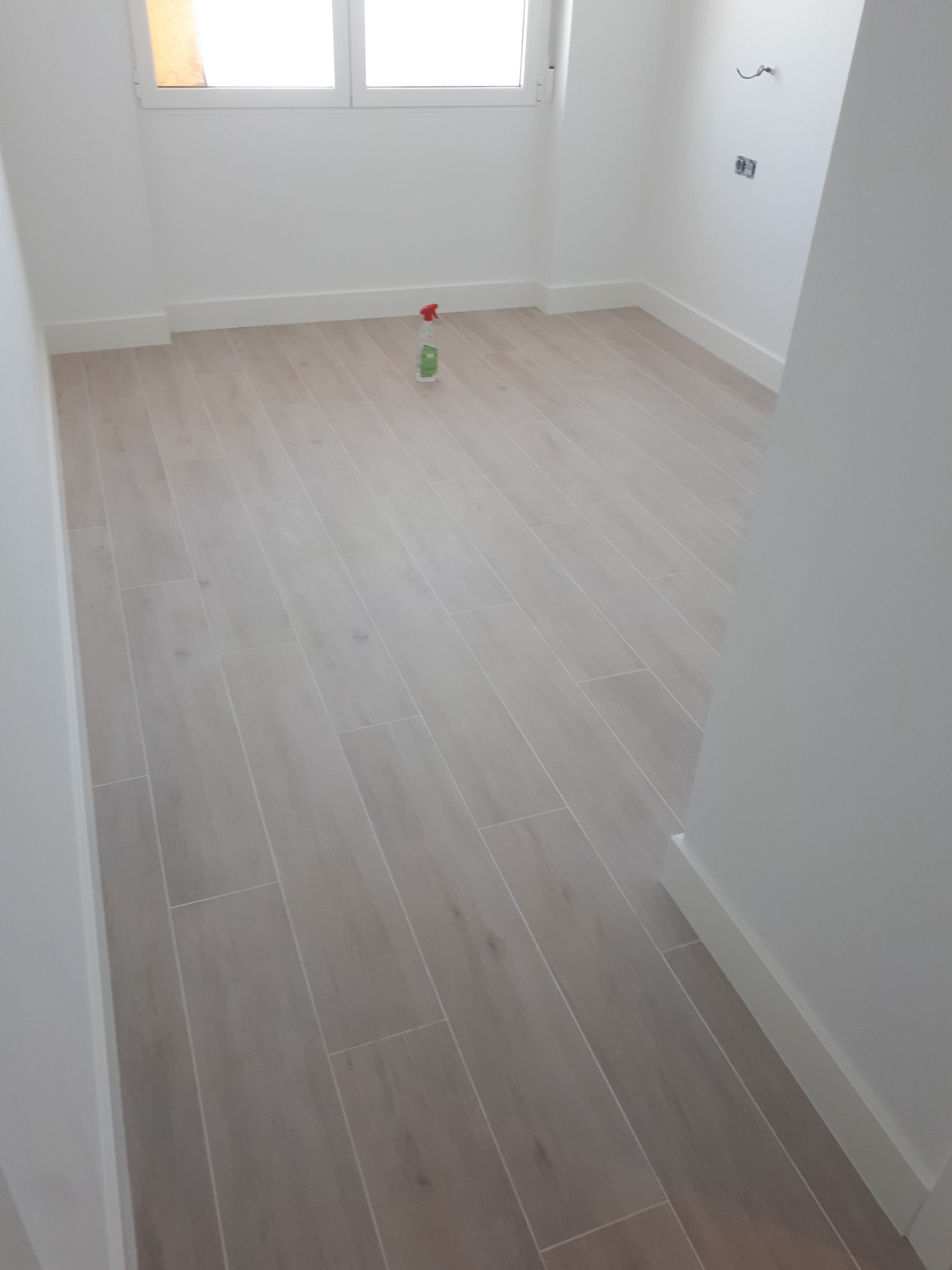 Jasne, podłużne płytki podłogowe ułożone w nowym, pustym pomieszczeniu z białymi ścianami i listwami przypodłogowymi, widoczne gniazdko elektryczne i butelka środka czyszczącego.