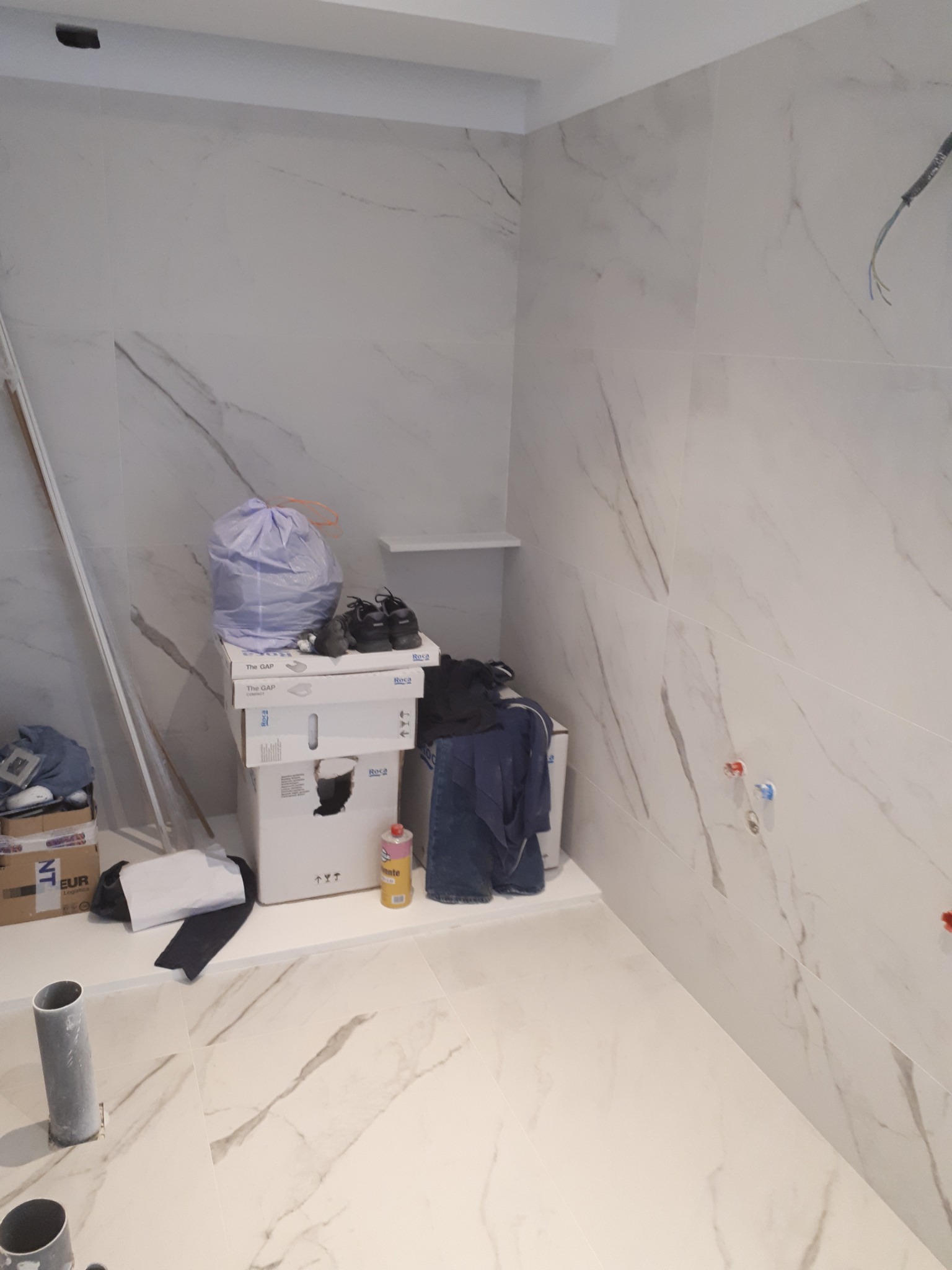 Wnętrze łazienki w trakcie prac, widoczne płytki imitujące marmur na ścianach i podłodze, wystające kable, rury, kartony z wyposażeniem i narzędzia.