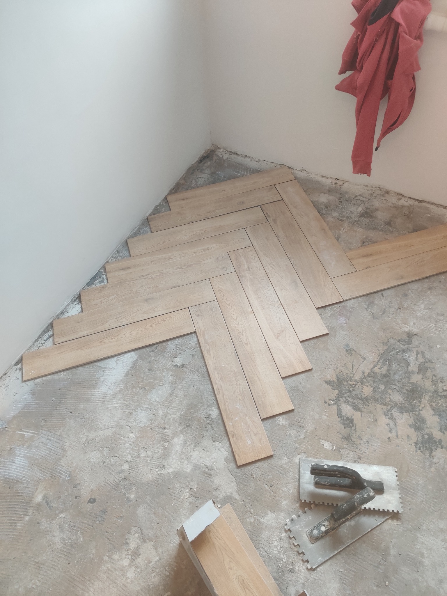 Układanie jasnych płytek podłogowych w jodełkę w rogu pomieszczenia, widoczne narzędzia glazurnicze i czerwona bluza wisząca na ścianie.