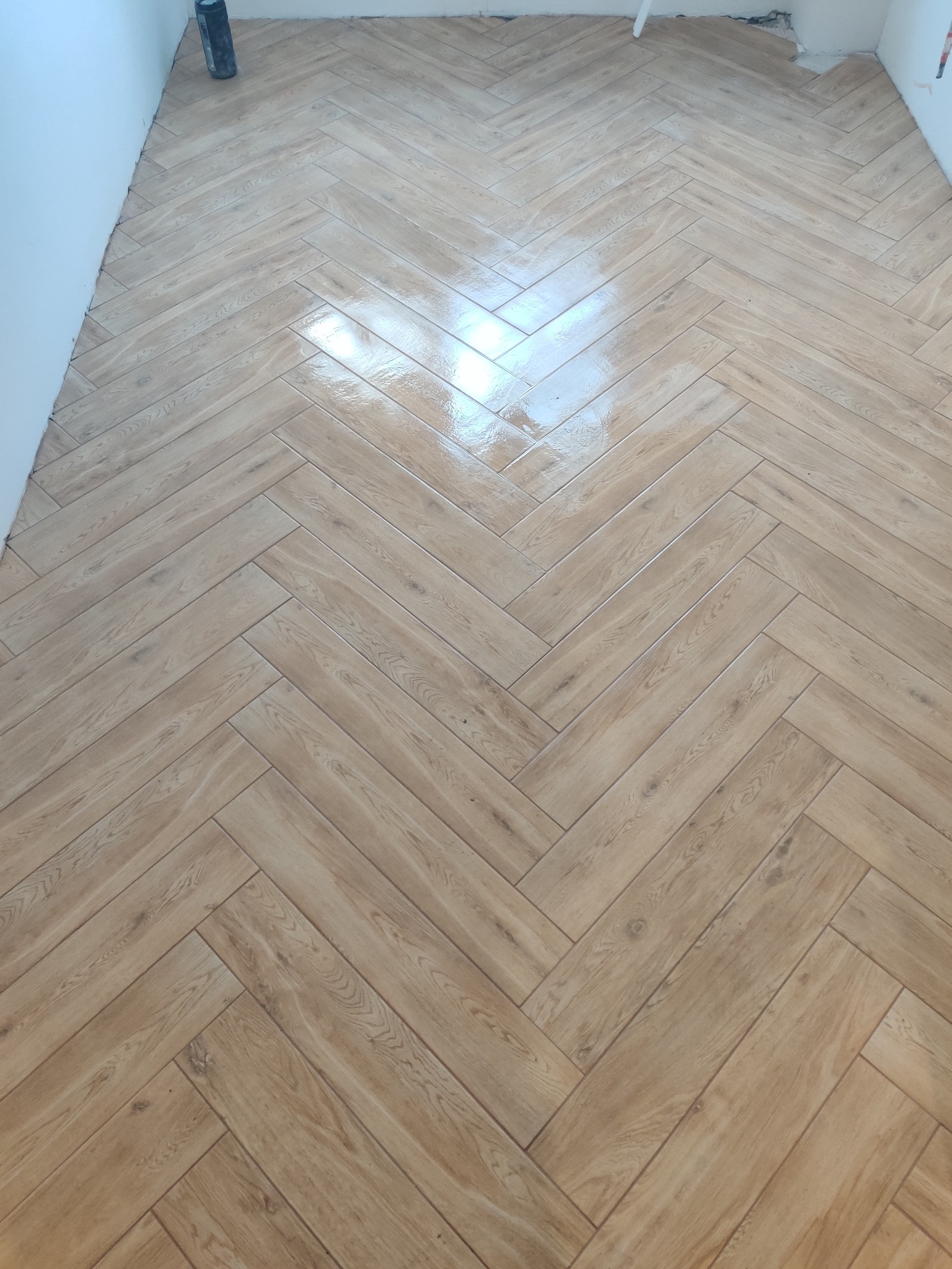 Ułożona w jodełkę, błyszcząca podłoga z płytek imitujących drewno, widoczny fragment ściany i rury w rogu.