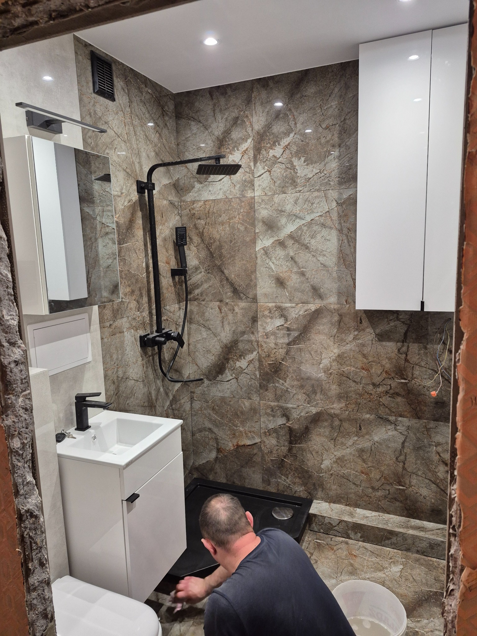 Nowoczesna łazienka z marmurowymi płytkami w odcieniach brązu i beżu, czarna armatura i białe meble łazienkowe, widoczna osoba czyszcząca podłogę przy kabinie prysznicowej