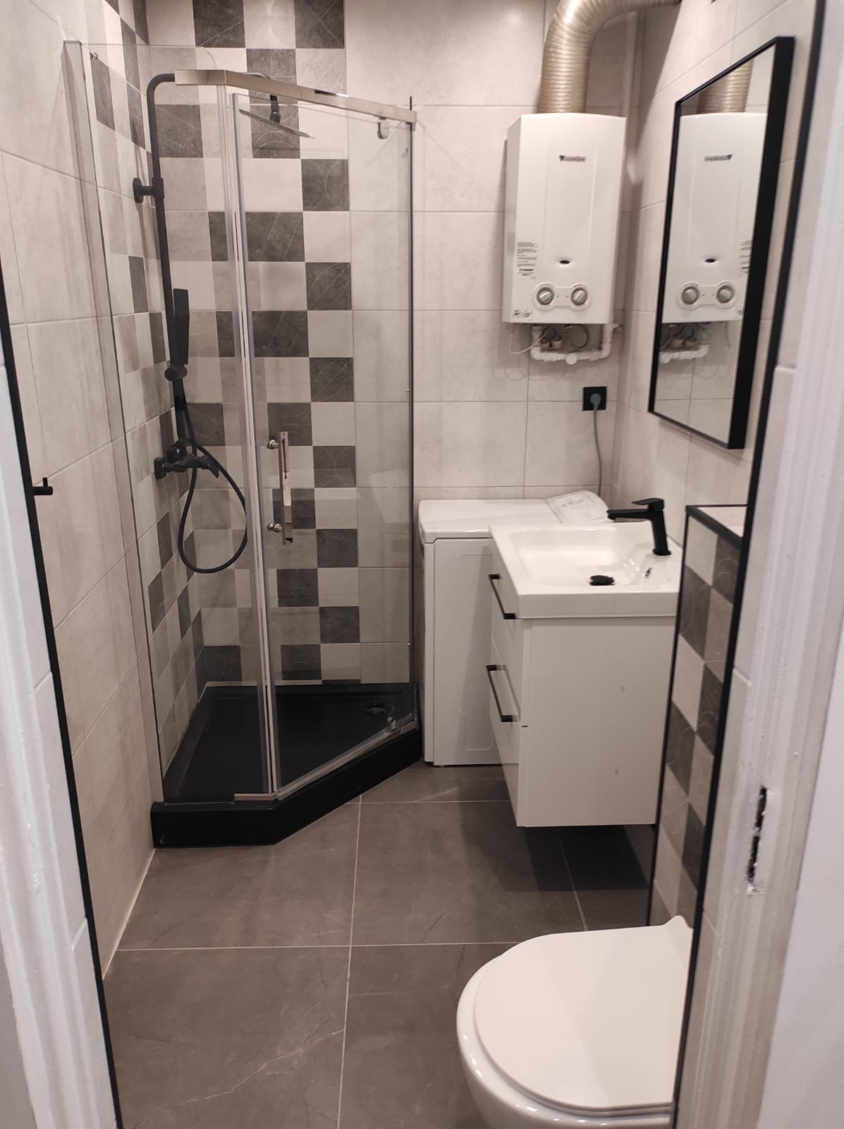 Wykończona łazienka z kabiną prysznicową z czarnym brodzikiem, białą szafką z umywalką i czarną armaturą, oraz grzejnikiem przepływowym i pralką w tle.